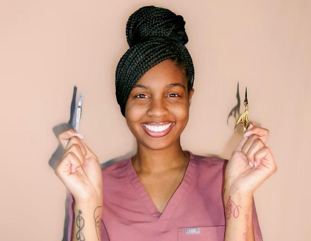 An S.B Esthetics esthetician in pink scrubs holding a dermaplaning blade and brow scissors.